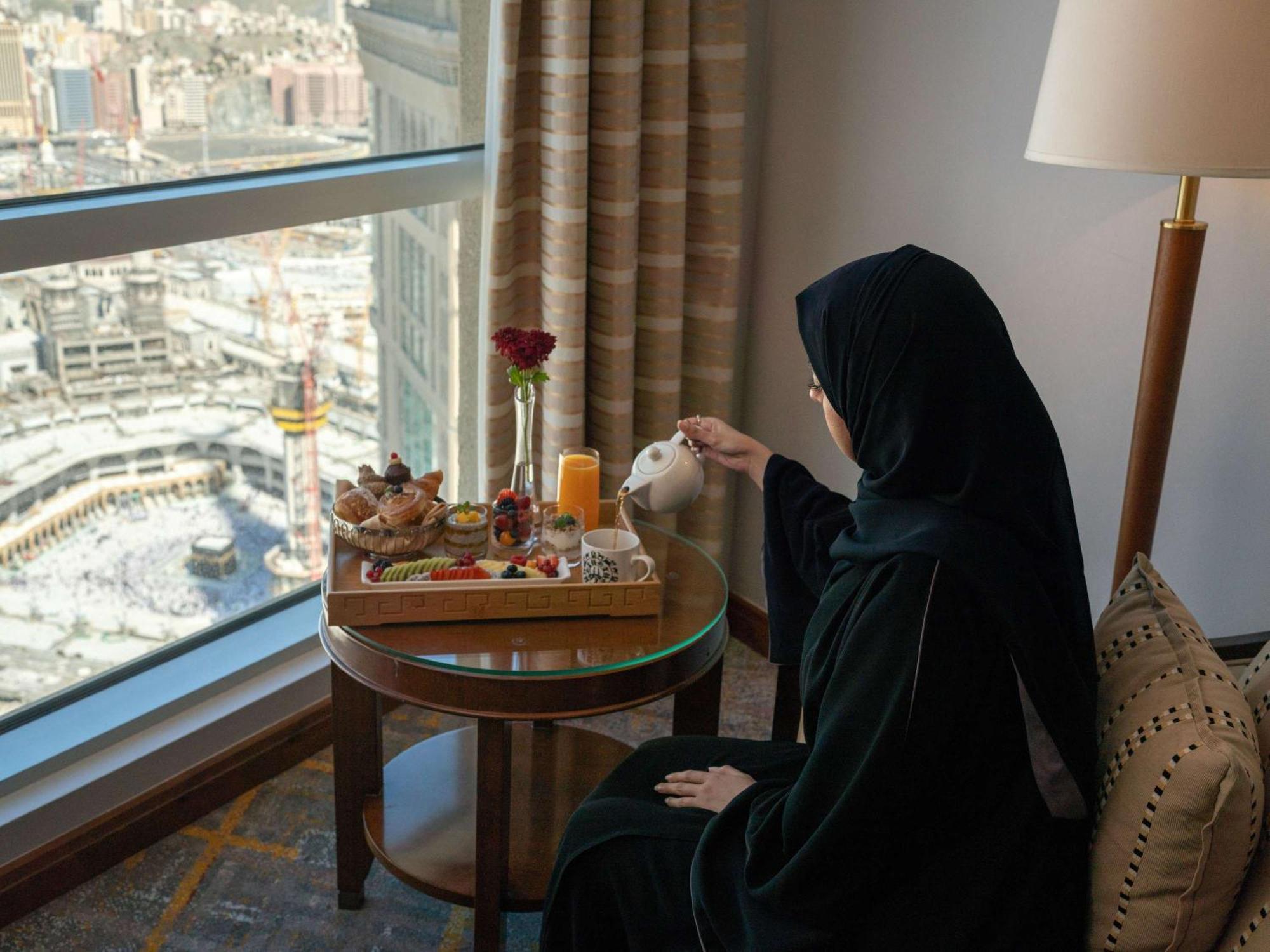 Swissotel Al Maqam Makkah Mekka Eksteriør billede The image depicts a woman wearing a black abaya, sitting at a table near a large window. She is pouring tea from a teapot into a cup. The table has a beautifully arranged tray with various food items, including pastries and fruits, as well as a glass