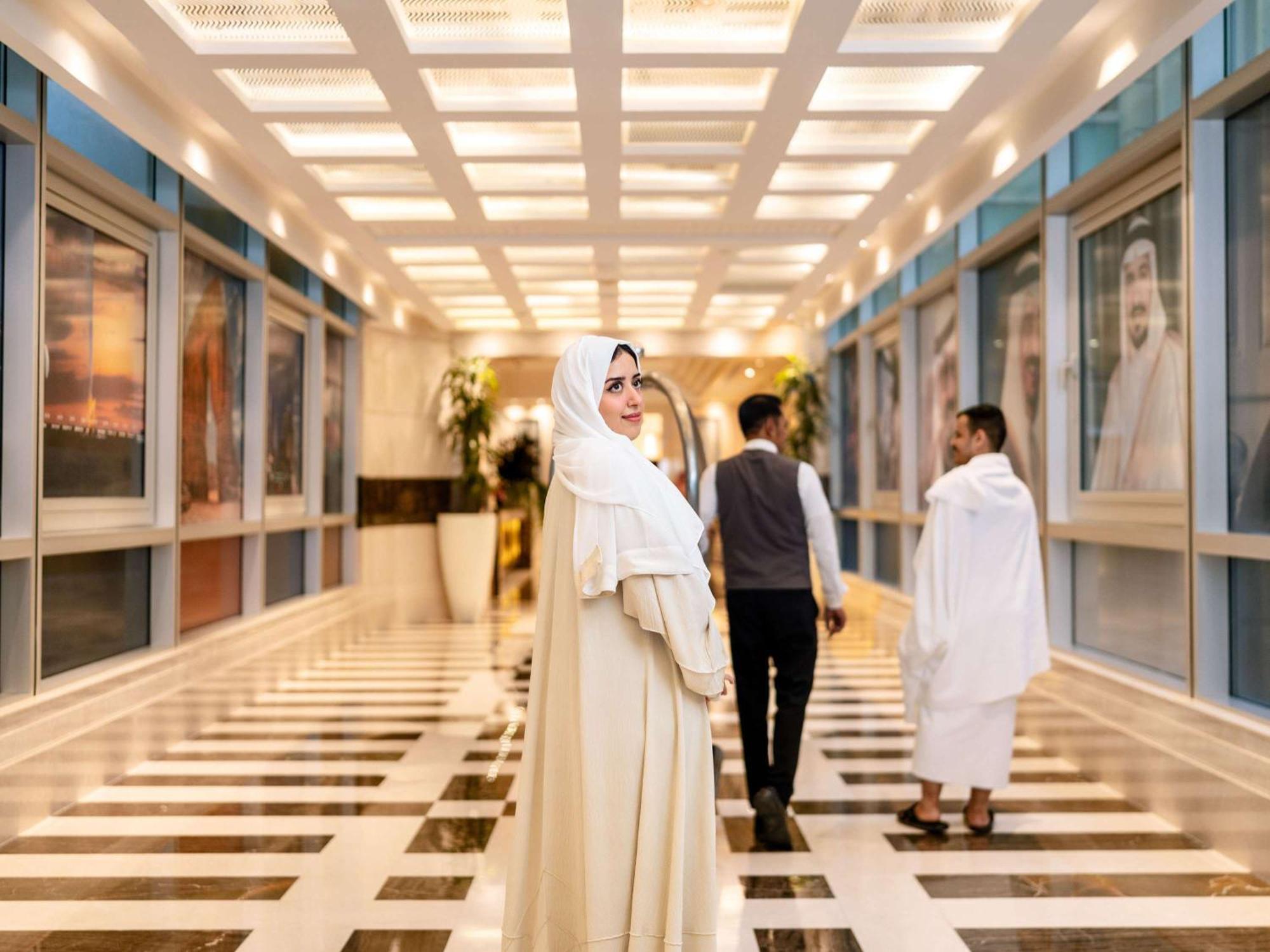 Swissotel Al Maqam Makkah Mekka Eksteriør billede The photo depicts a modern corridor with a shiny tiled floor and bright overhead lighting. In the foreground, a woman wearing a long white dress and a headscarf is looking back at the camera. She appears to be confident and stylish. Two other individ