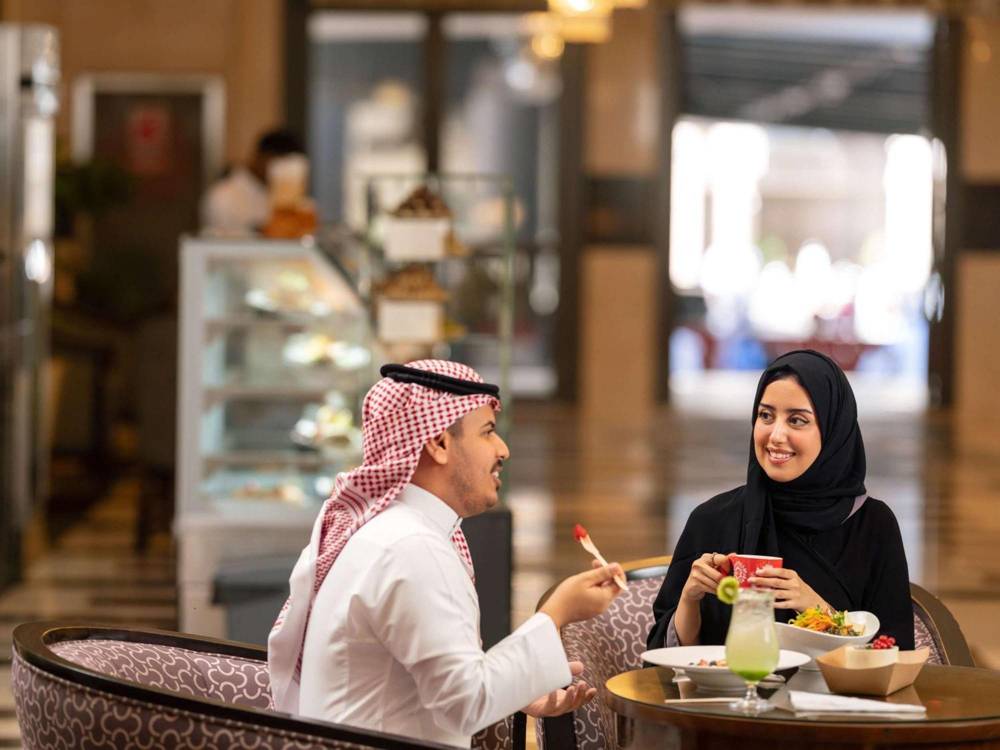 Swissotel Al Maqam Makkah Mekka Eksteriør billede The photo features a man and a woman engaged in a conversation at a dining table. The man is wearing traditional attire with a white thobe and a headscarf, while the woman is dressed in a black abaya and headscarf. They appear to be enjoying a meal t