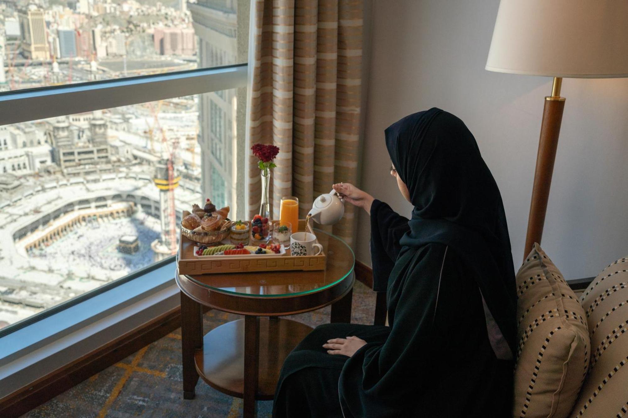 Swissotel Al Maqam Makkah Mekka Eksteriør billede The photo shows a woman sitting at a table by a large window in a hotel room. She is wearing a black abaya and is pouring tea from a teapot into a cup. On the table in front of her, there is a tray filled with various food items, including pastries a