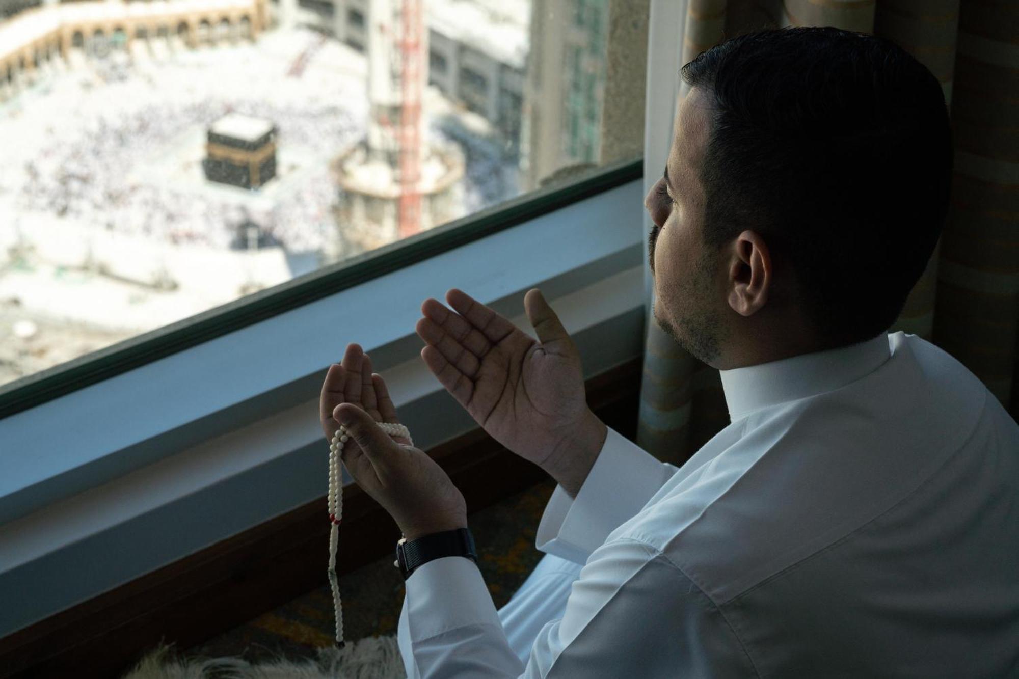 Swissotel Al Maqam Makkah Mekka Eksteriør billede The photo shows a man sitting next to a window, engaged in prayer. He is facing toward the direction of the Kaaba, which is visible in the background, suggesting he is in Mecca. The man is holding prayer beads in one hand while gesturing in supplicat