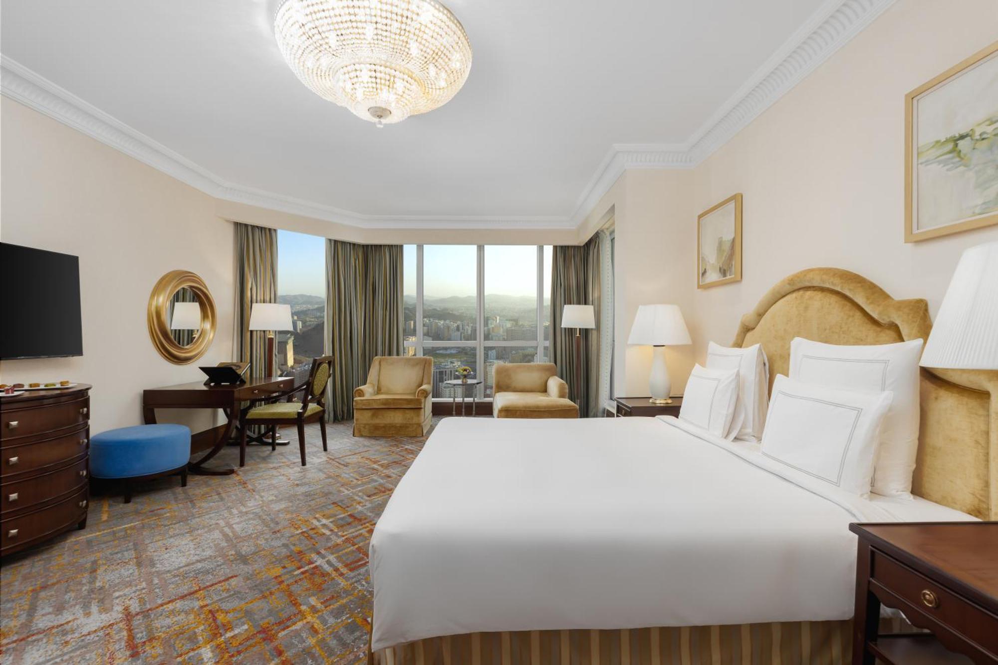 Swissotel Al Maqam Makkah Mekka Eksteriør billede The photo shows a well-furnished hotel room with a modern and elegant design. It features a large bed with white linens and decorative pillows. There are two armchairs nearby, a small round table, and a writing desk with a chair. A television is moun