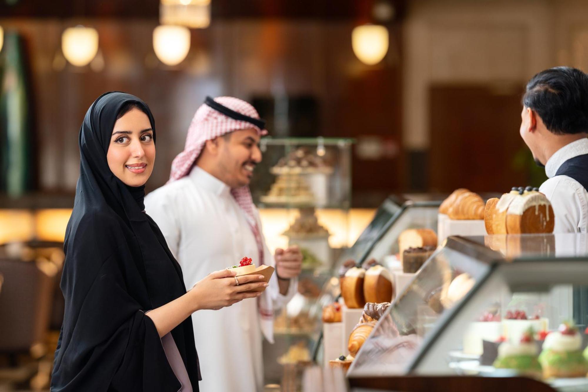 Swissotel Al Maqam Makkah Mekka Eksteriør billede The photo shows a scene in a pastry shop or dessert counter. A woman dressed in a black abaya is holding a dessert and smiling at the camera. In the background, a man wearing traditional Middle Eastern attire is interacting with another staff member 
