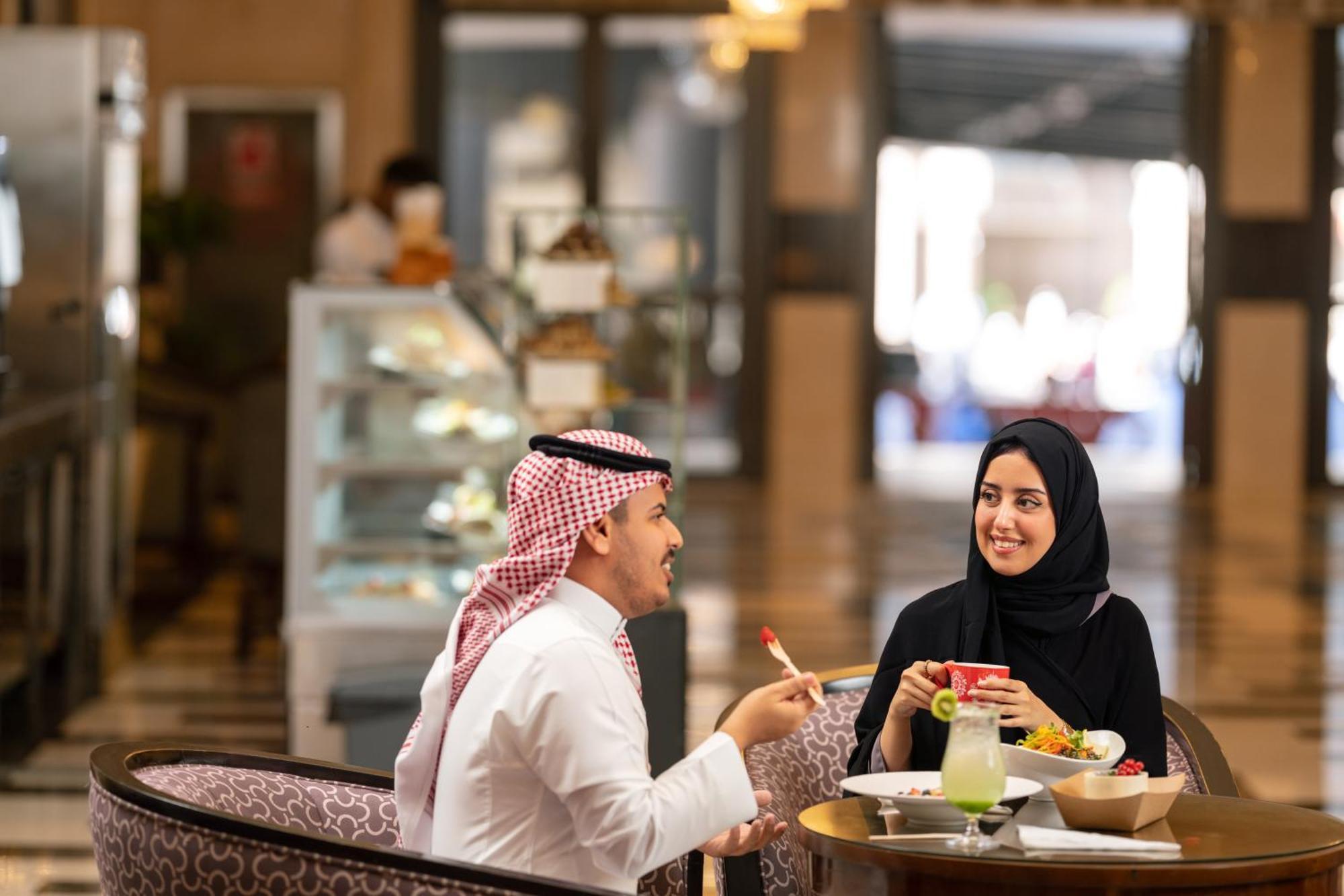 Swissotel Al Maqam Makkah Mekka Eksteriør billede The photo depicts a cheerful scene in a cafe or restaurant setting. A man, dressed in traditional attire with a headscarf, is engaged in a friendly conversation with a woman who is wearing a black hijab. They are seated at a table, which has several 