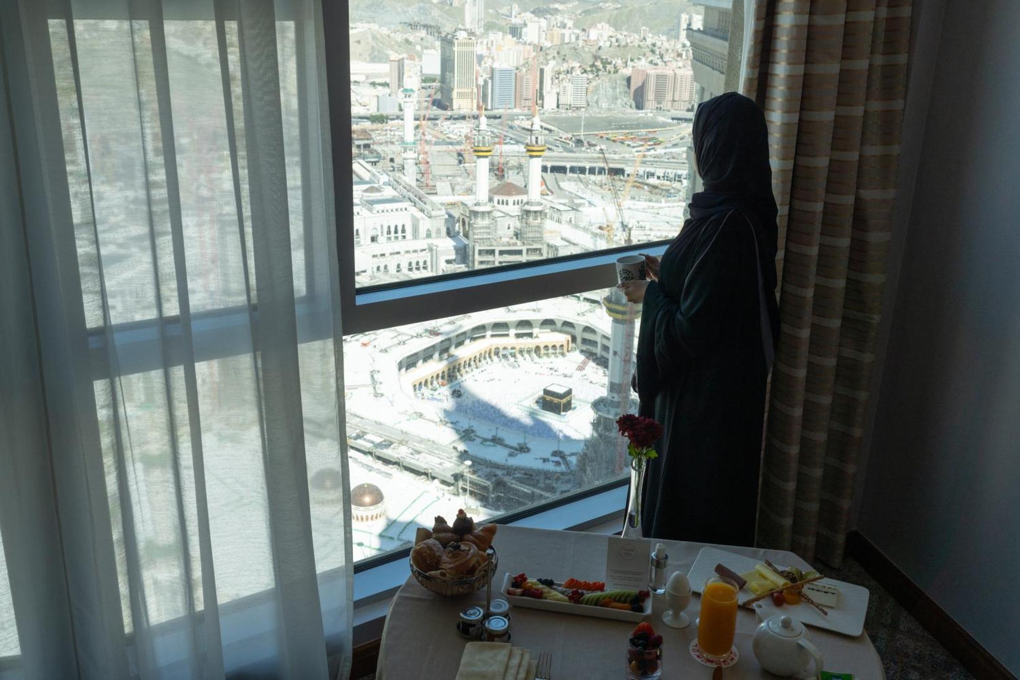 Swissotel Al Maqam Makkah Mekka Eksteriør billede The photo depicts a woman standing by a large window, gazing out at a cityscape. She is wearing a dark, traditional outfit and appears to be in a hotel room. In front of her, on a table, there is a spread of breakfast items, including fruits and drin