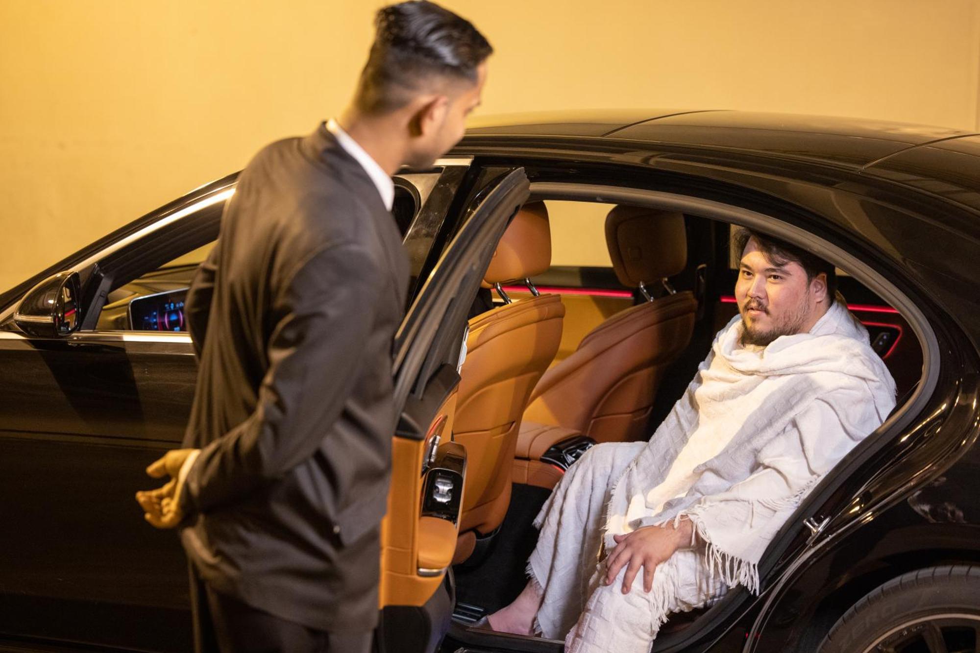 Swissotel Al Maqam Makkah Mekka Eksteriør billede The image shows a scene outside a car at night. One person, dressed in a formal black suit, is standing next to an open car door, seemingly talking to the other person inside the car. The person inside the car appears to be wearing a white blanket or