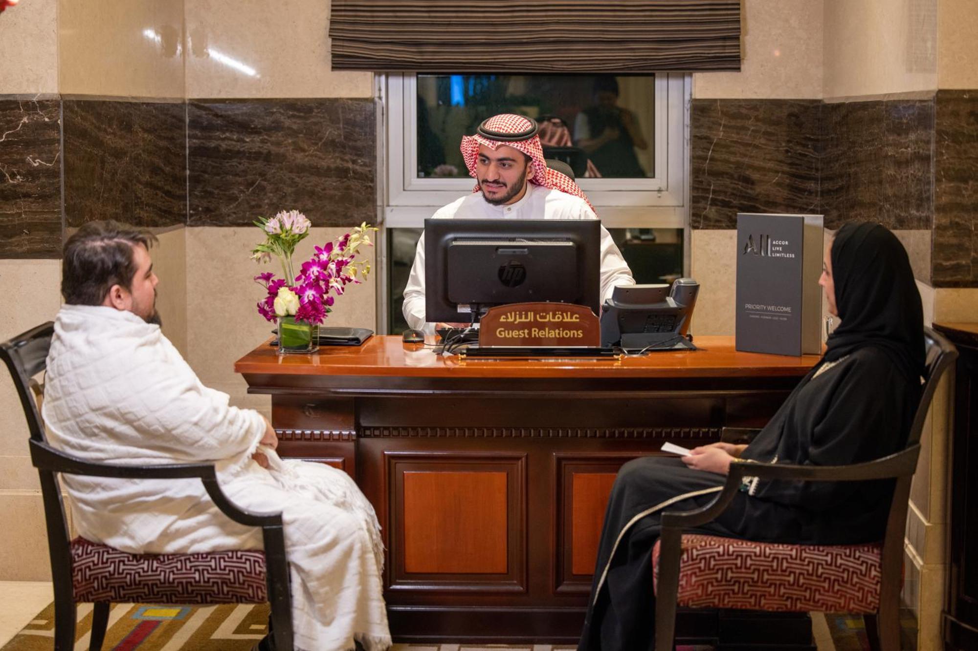 Swissotel Al Maqam Makkah Mekka Eksteriør billede The photo shows a reception area with a male receptionist seated at a desk, wearing traditional attire, including a headscarf. He is focused on a computer screen. In front of him, there are two individuals seated: one is a man in traditional clothing