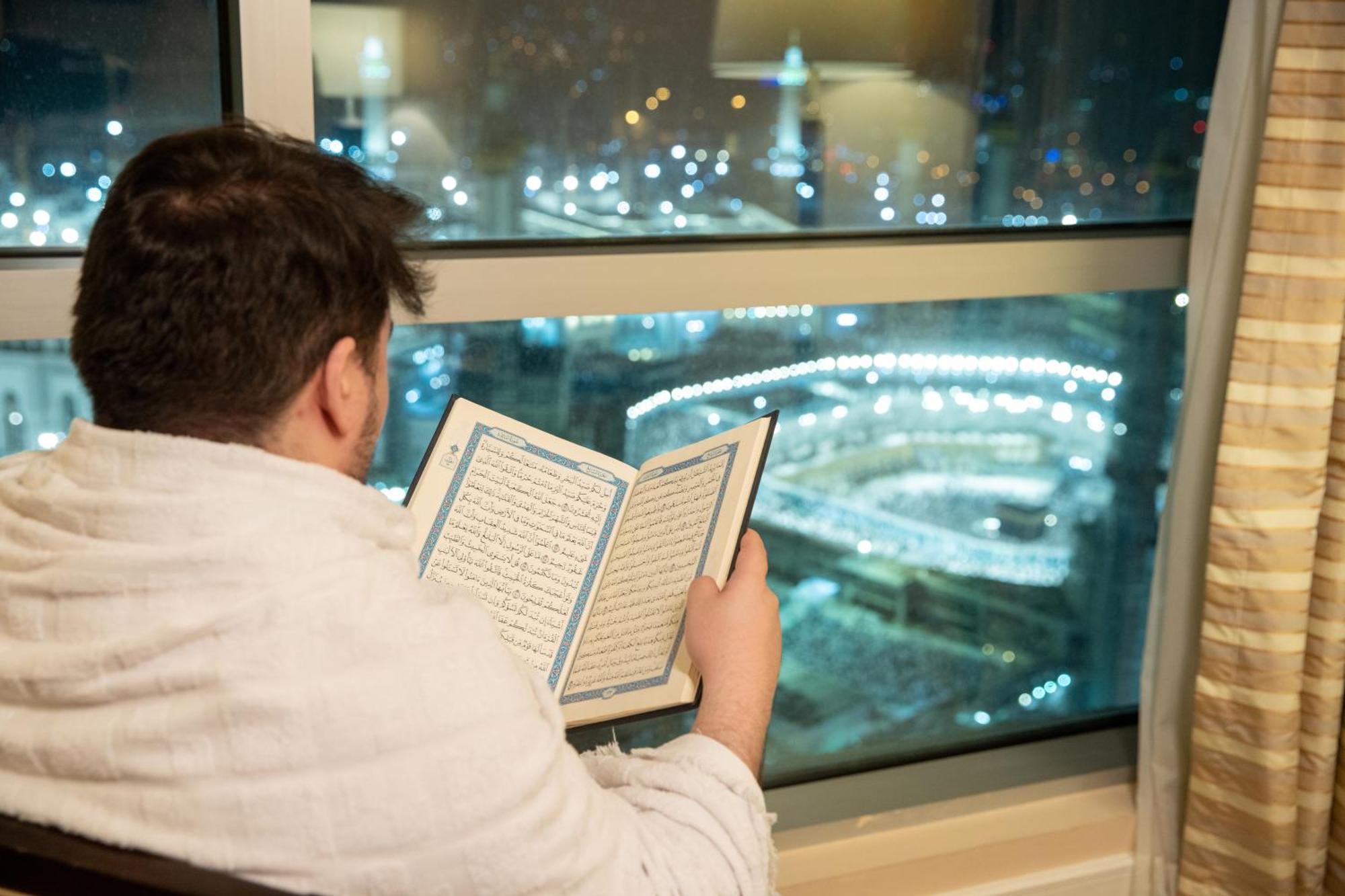 Swissotel Al Maqam Makkah Mekka Eksteriør billede The photo shows a person sitting by a window, reading a book while looking out at a nighttime cityscape. The view outside features bright lights, suggesting a bustling environment, possibly a significant religious site. The individual appears to be w