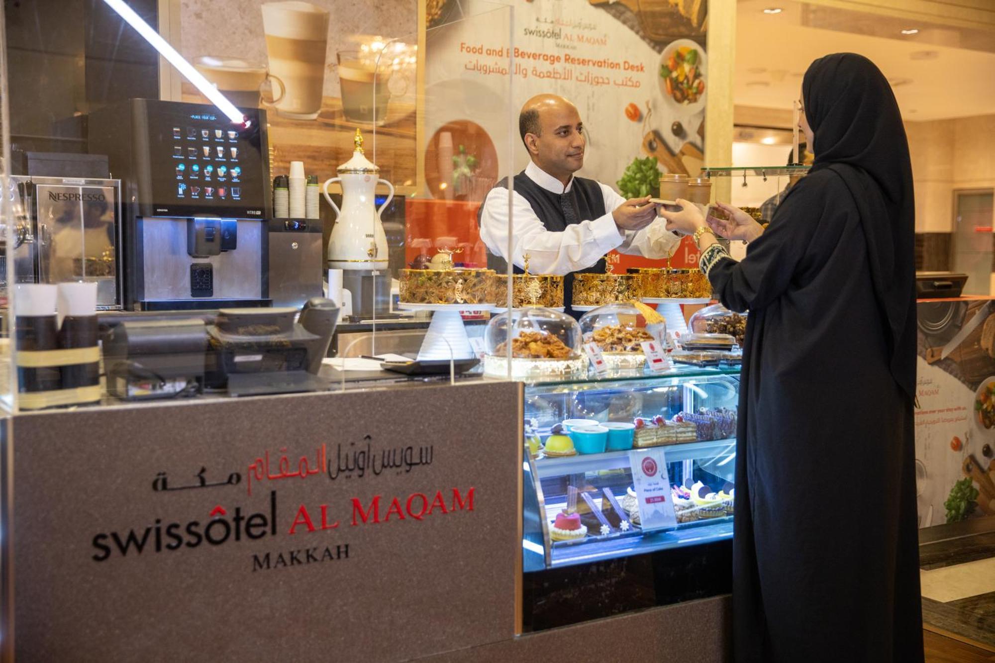 Swissotel Al Maqam Makkah Mekka Eksteriør billede The photo shows an interaction at a café or restaurant located in the Swissôtel Al Maqam, Makkah. On the left side, there is a display counter filled with various pastries and desserts. A male staff member, dressed in a white shirt and black vest, is