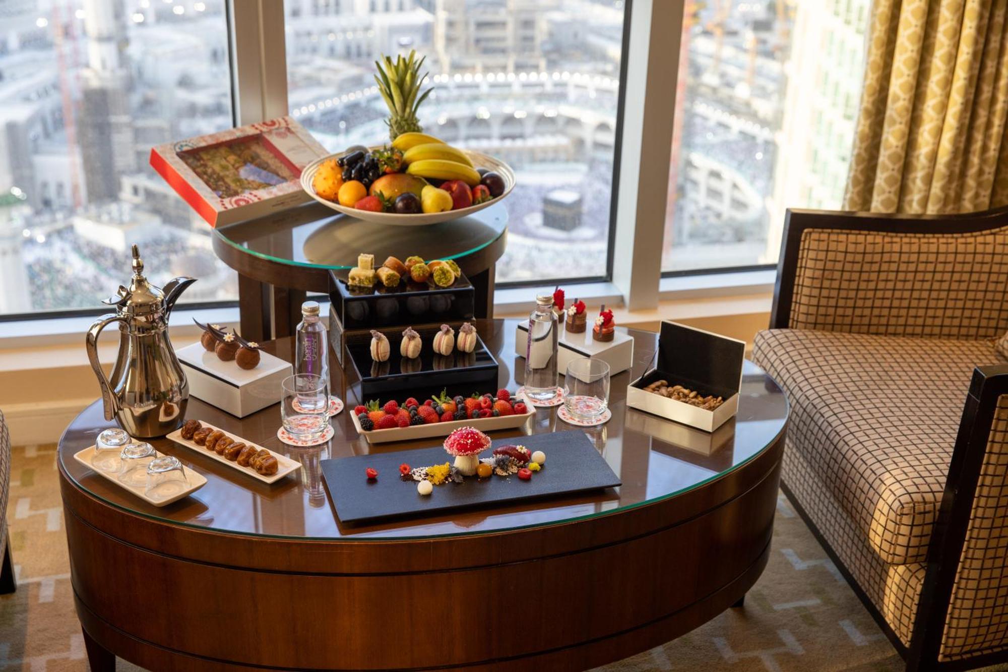 Swissotel Al Maqam Makkah Mekka Eksteriør billede The photo shows a lavish spread of refreshments arranged on a table in a hotel room with a view. There is a large bowl of fresh fruits, including bananas, at the center. Surrounding the fruit bowl are various desserts, including chocolate-dipped item