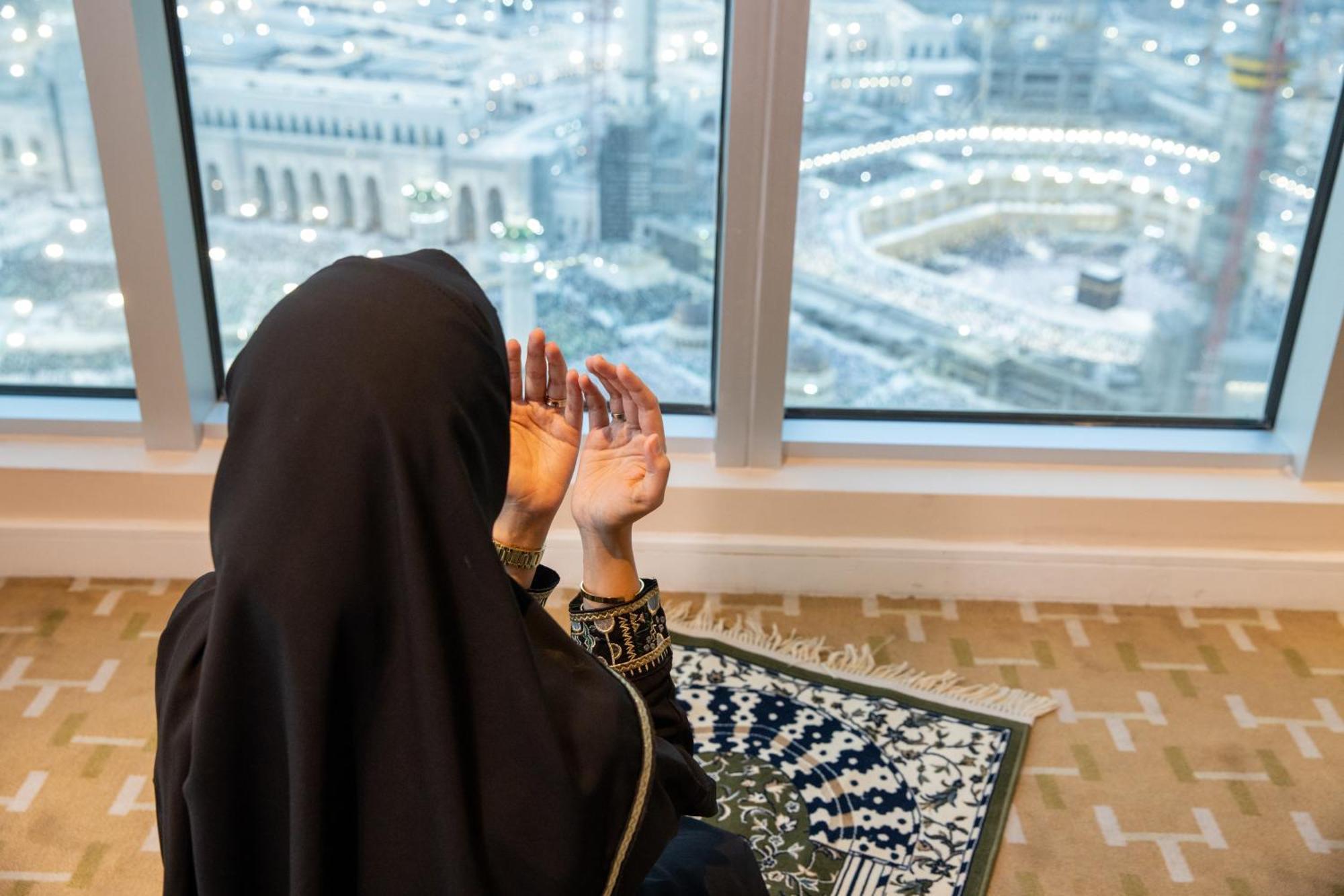 Swissotel Al Maqam Makkah Mekka Eksteriør billede The image shows a woman in a black hijab sitting on a prayer mat, facing a large window that overlooks a cityscape. She is engaged in prayer, with her hands raised in a gesture of supplication. The view reveals a significant location, likely a religi