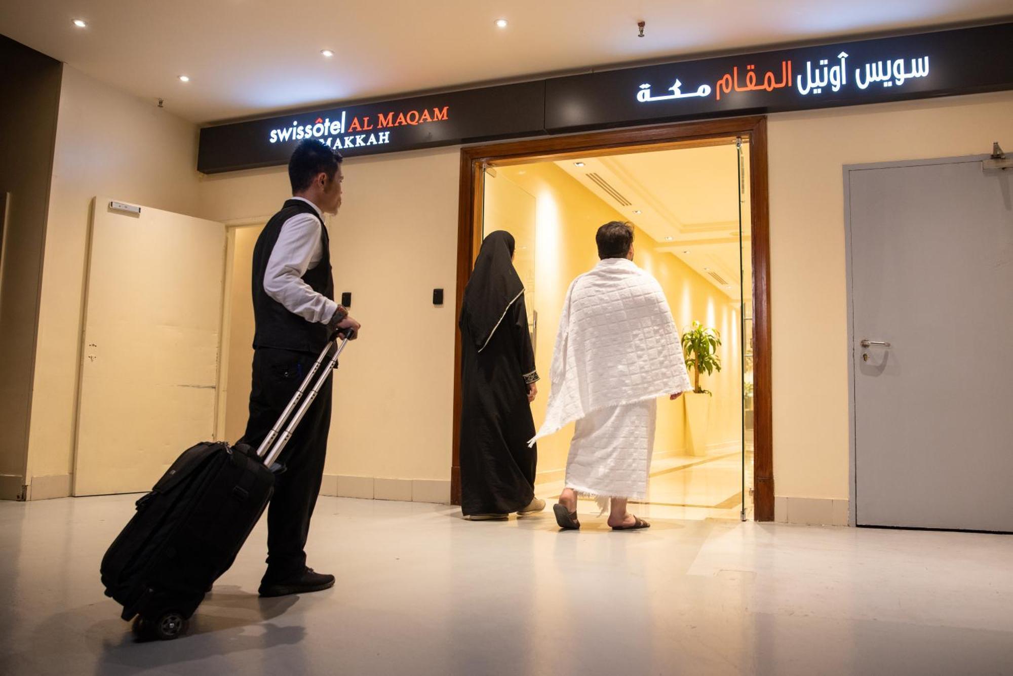 Swissotel Al Maqam Makkah Mekka Eksteriør billede The photo shows a hotel entrance, specifically for the "Swissotel Al Maqam Makkah," as indicated by the sign. In the scene, a man in a formal outfit carrying a suitcase waits nearby, while two individuals, one in traditional clothing (possibly a woma