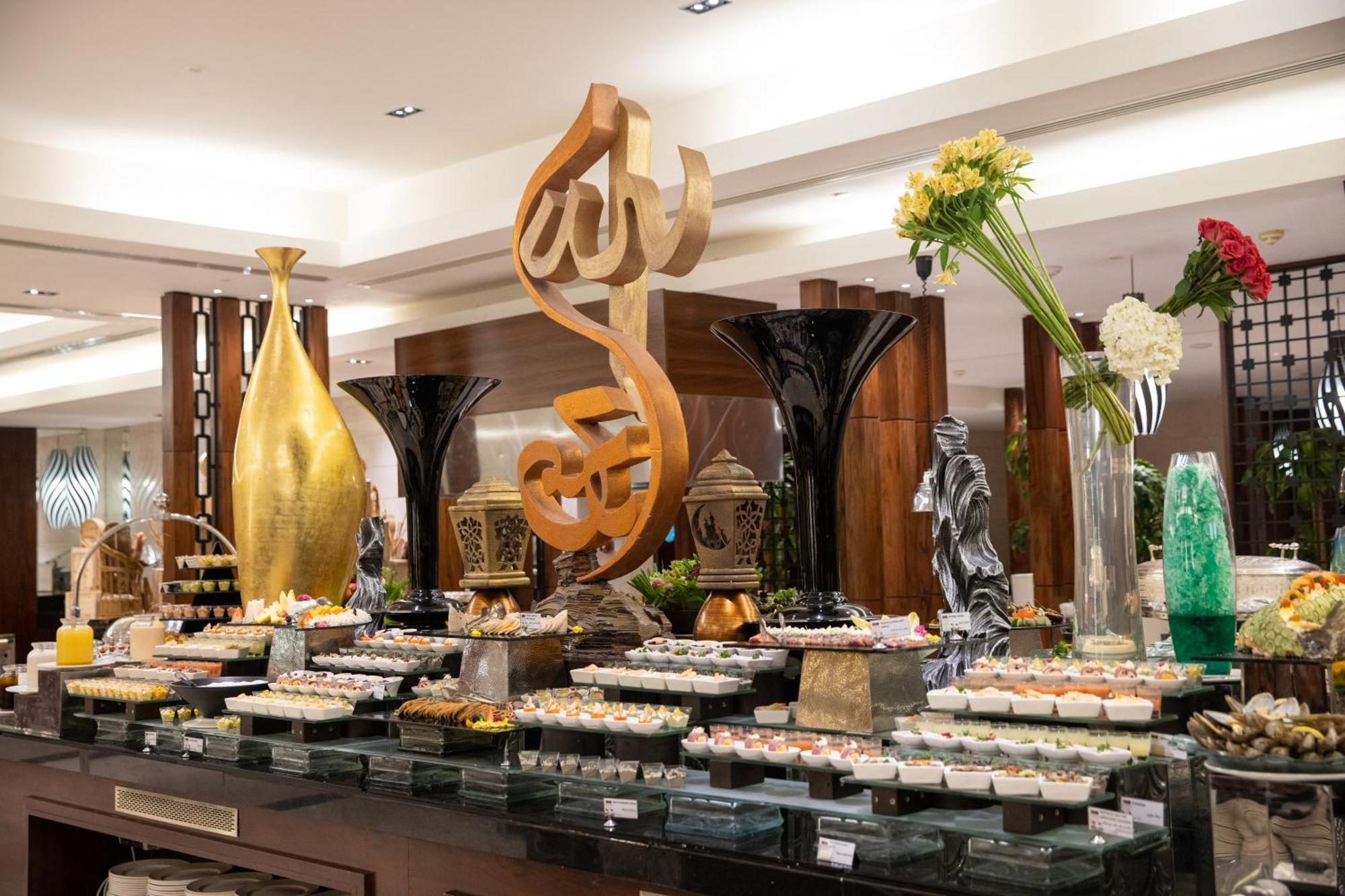 Swissotel Al Maqam Makkah Mekka Eksteriør billede The photo displays a beautifully arranged buffet set up in a restaurant or hotel. In the center, there is an artistic sculpture featuring a stylized calligraphic design. Surrounding it are various decorative elements, including vases and floral arran