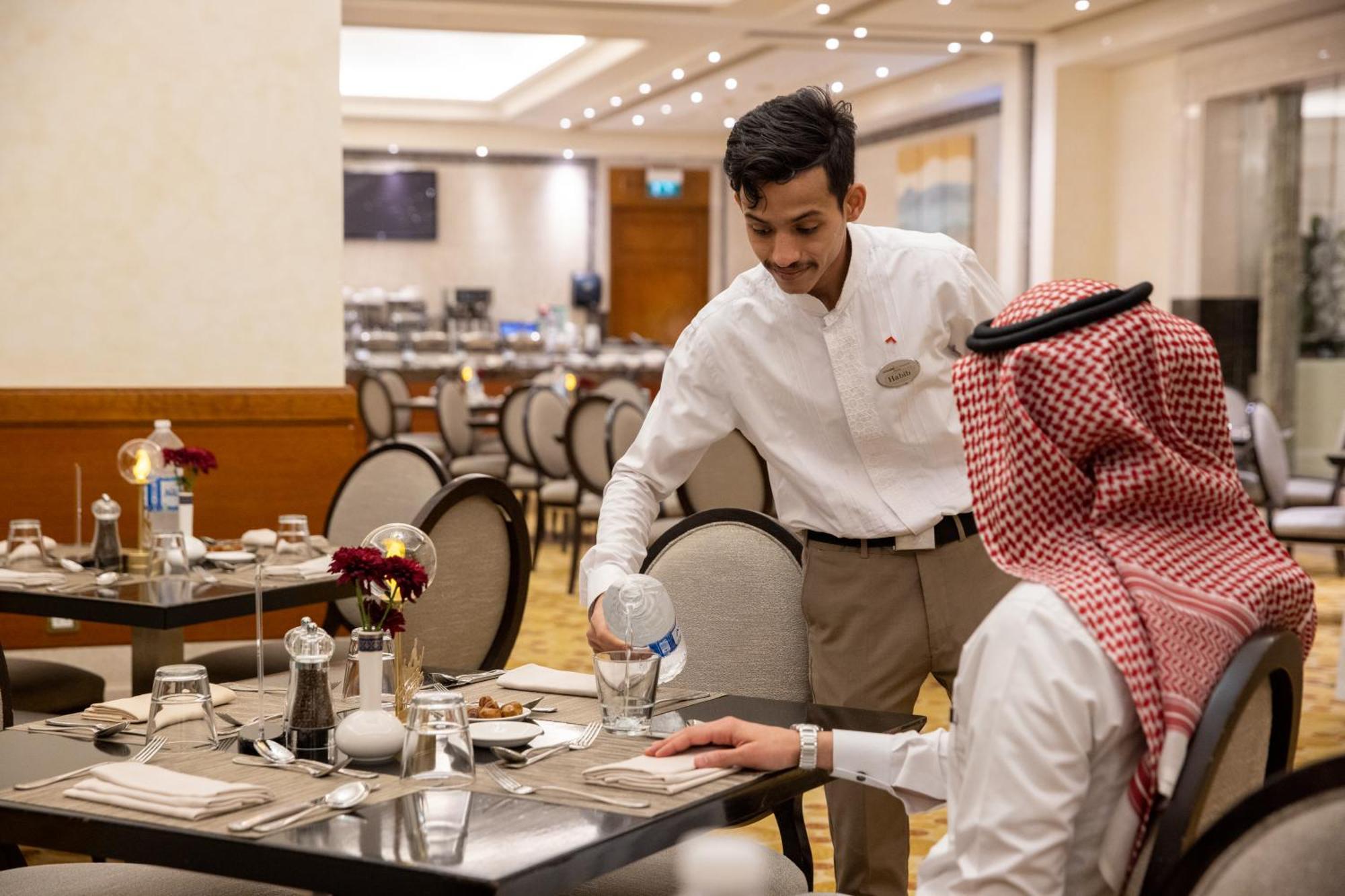 Swissotel Al Maqam Makkah Mekka Eksteriør billede The photo shows a dining scene in a restaurant. A waiter, dressed in a white shirt and light-colored pants, is pouring water into a glass placed on the table. The table is set with plates, cutlery, and a small vase with flowers. The diner, who is wea