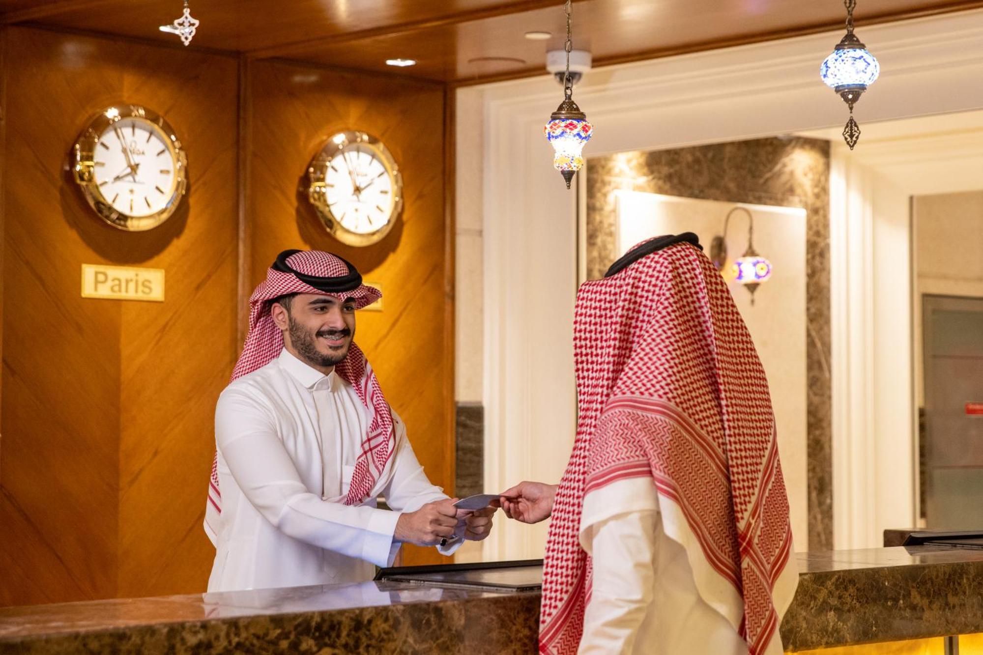Swissotel Al Maqam Makkah Mekka Eksteriør billede The photo shows a hotel reception area. In the foreground, a staff member is interacting with a guest. The staff member, dressed in traditional attire, appears to be handing something to the guest. Behind the reception desk, there are several wall cl