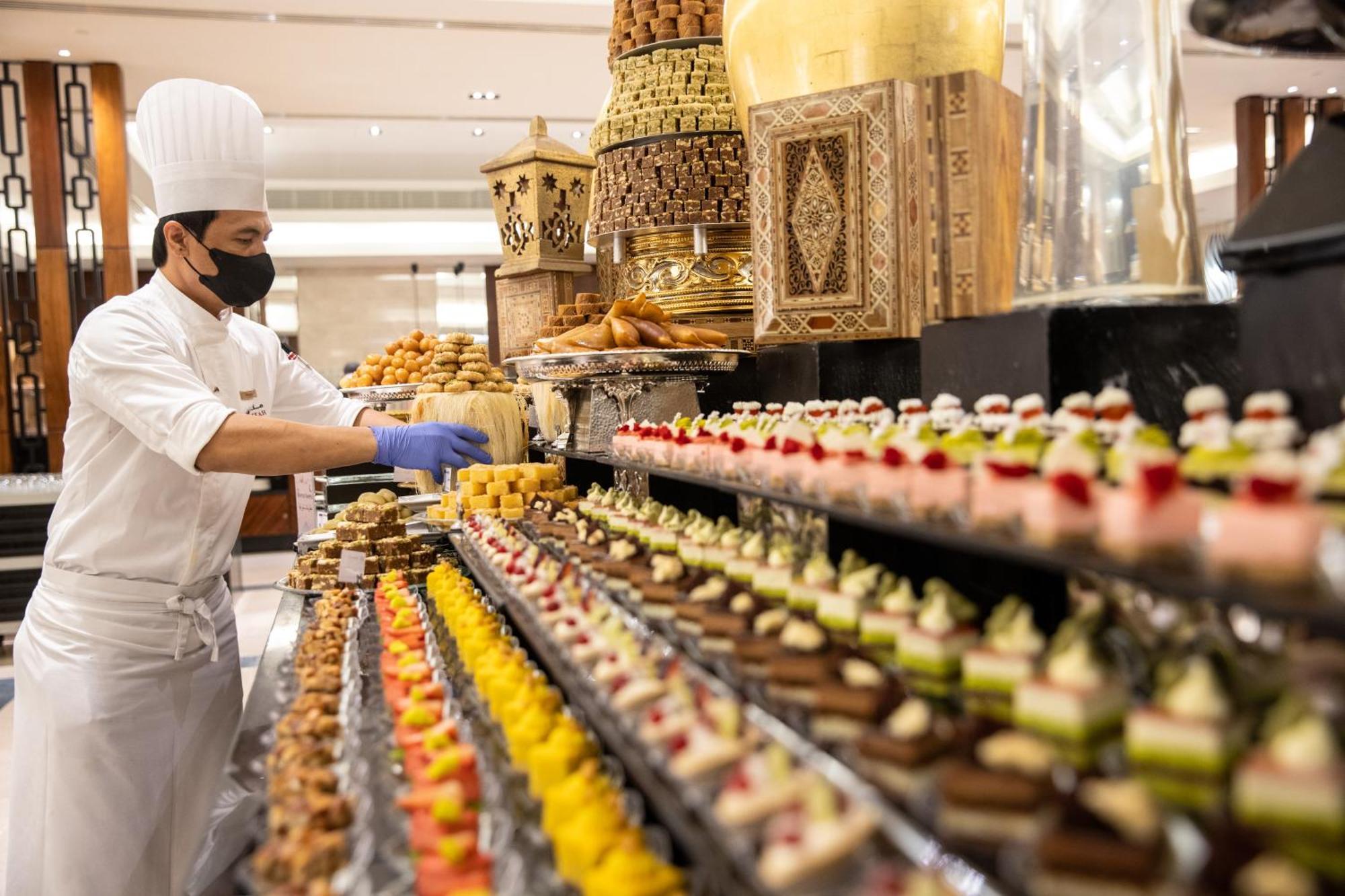 Swissotel Al Maqam Makkah Mekka Eksteriør billede The photo depicts a chef wearing a white uniform and a traditional chef's hat, working in a setting that appears to be a buffet or dessert display area. The chef is carefully arranging a variety of colorful pastries and desserts on long tables. The d
