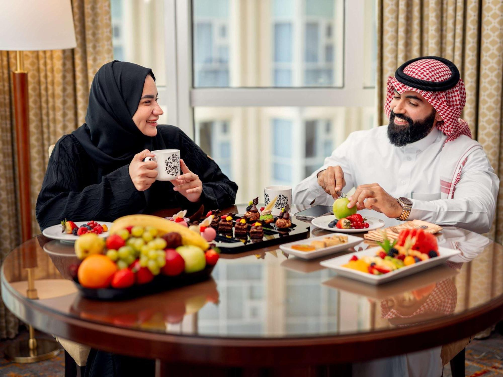 Swissotel Al Maqam Makkah Mekka Eksteriør billede The photo shows two people enjoying a meal together at a dining table. The woman is wearing a black abaya and smiling while holding a cup, and the man is dressed in traditional white attire with a red and white checkered headscarf. They are surrounde