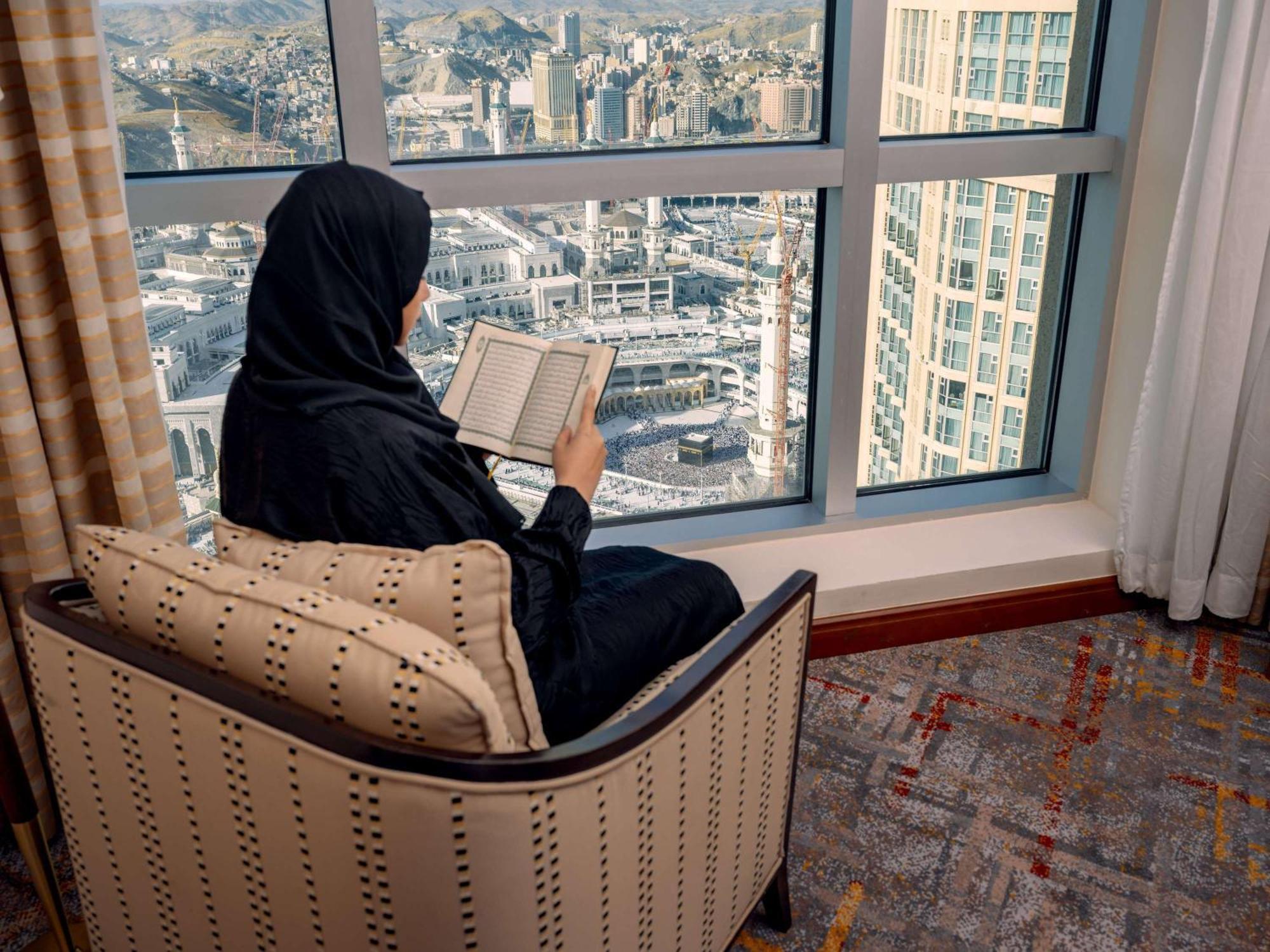 Swissotel Al Maqam Makkah Mekka Eksteriør billede The photo depicts a woman seated in a comfortable chair by a large window. She is wearing a traditional black garment and is focused on reading a book. Outside the window, there is a view of a cityscape, possibly a metropolitan area, with various bui