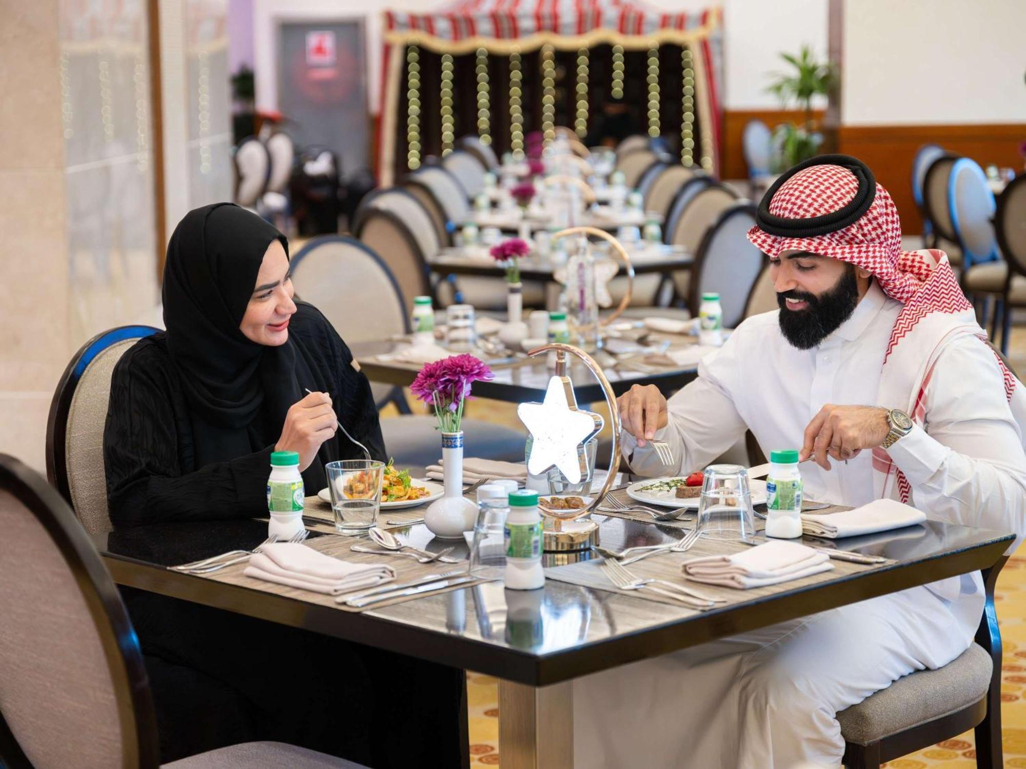 Swissotel Al Maqam Makkah Mekka Eksteriør billede The photo shows a couple dining at a restaurant. The man is wearing traditional attire, including a white thobe and a red and white checkered headscarf, while the woman is dressed in a black abaya and a headscarf. They are engaged in a pleasant conve