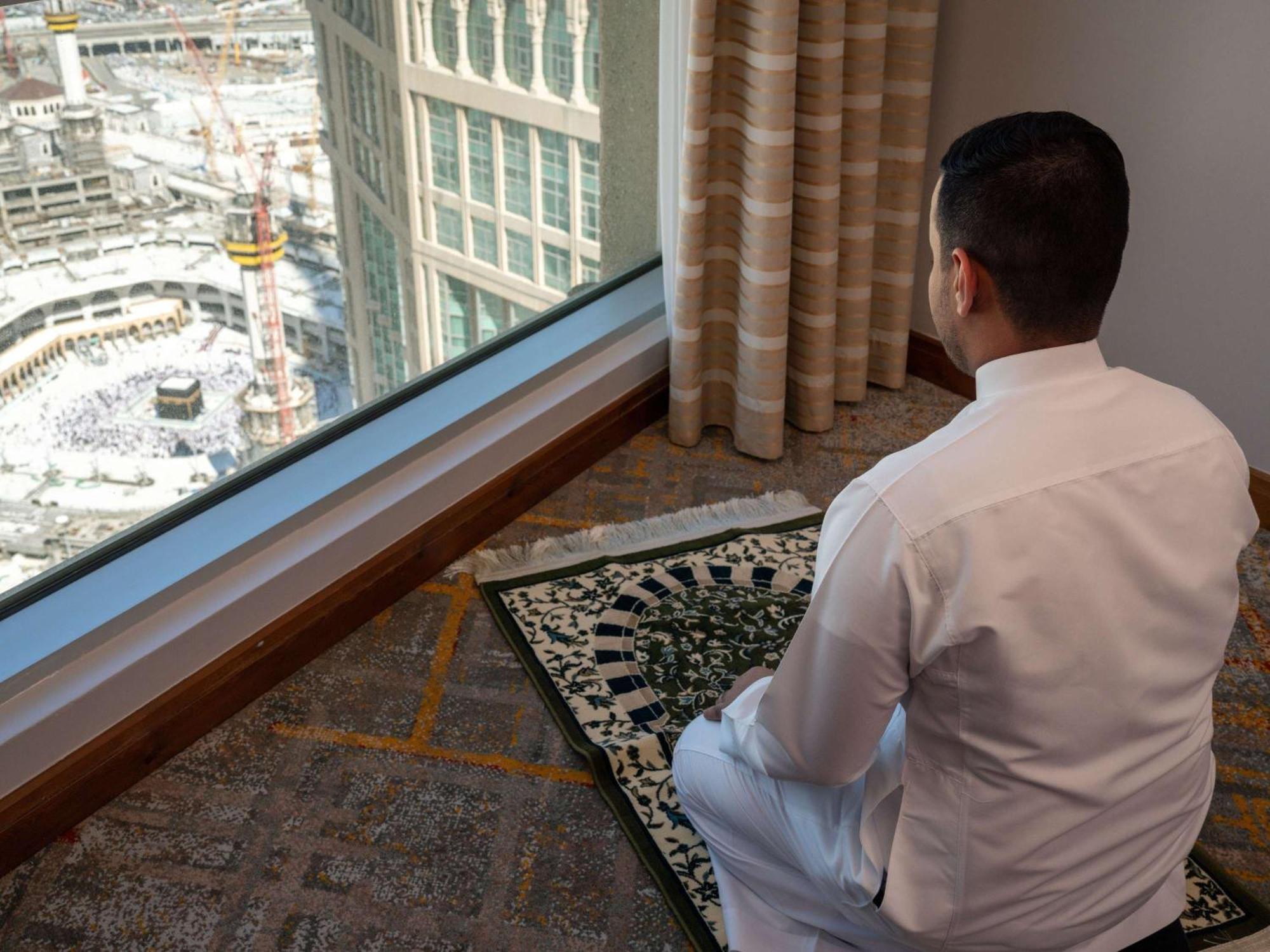 Swissotel Al Maqam Makkah Mekka Eksteriør billede The photo shows a man sitting in a prayer position on a prayer mat, facing a large window. Through the window, there's a view of a construction site, possibly near a significant religious site, given the context. The man is wearing traditional attire