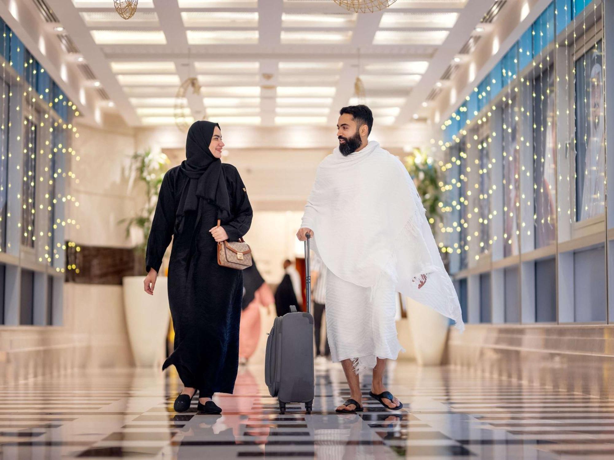 Swissotel Al Maqam Makkah Mekka Eksteriør billede The photo depicts a couple walking together in a modern, well-lit hallway. The man is dressed in traditional white garments, while the woman is wearing a black abaya. The hallway features a stylish marble floor and is adorned with decorative lights, 