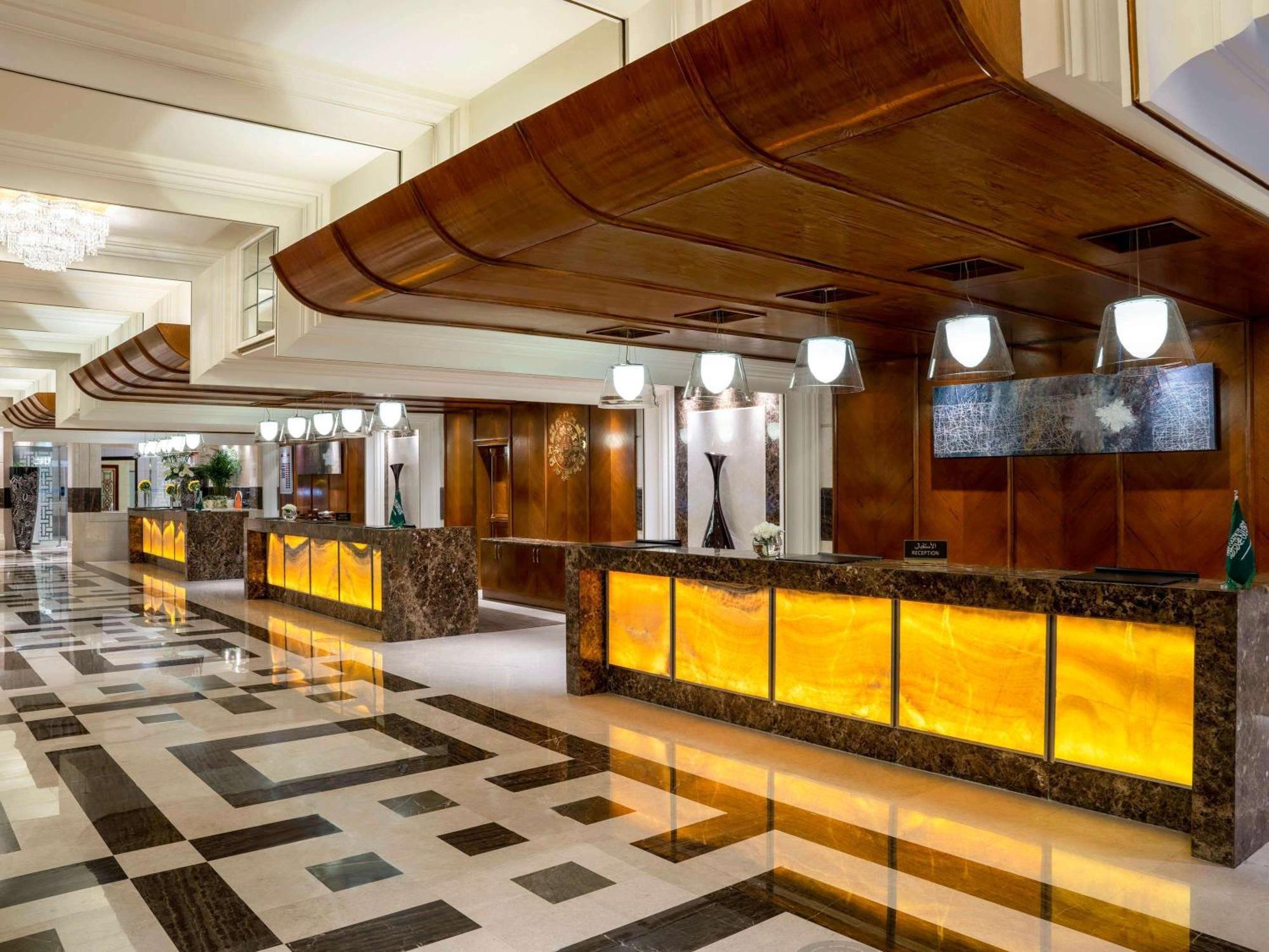 Swissotel Al Maqam Makkah Mekka Eksteriør billede The image shows a hotel lobby with an elegant and sophisticated design. There are multiple reception desks made of dark wood and marble, featuring backlit panels that emit a warm, glowing light. The ceiling has a unique curved wooden structure, addin