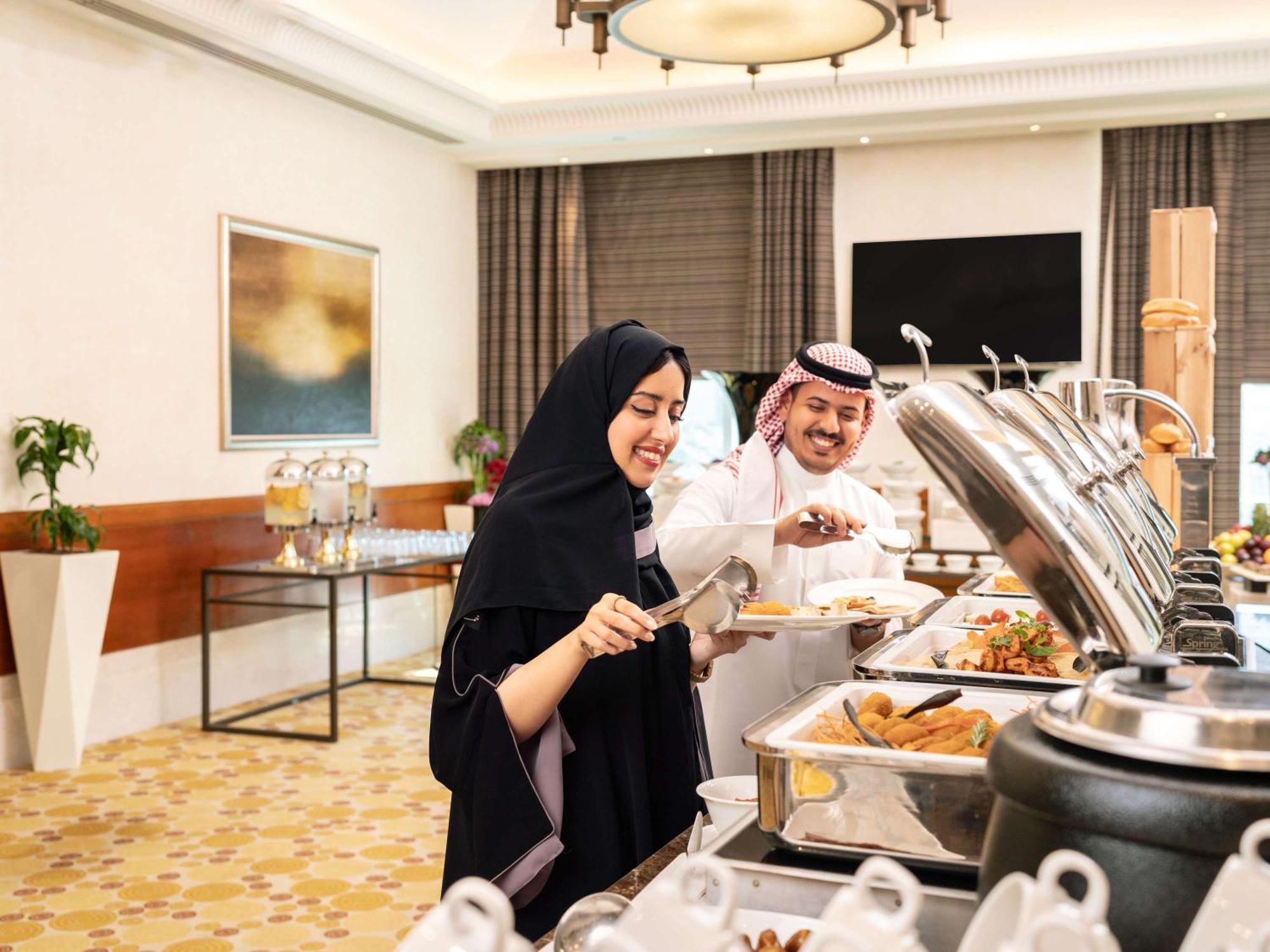 Swissotel Al Maqam Makkah Mekka Eksteriør billede The photo depicts a buffet setting where two people are serving food. A woman wearing a traditional black abaya is smiling as she uses tongs to select food from a buffet tray. Next to her, a man dressed in traditional attire, including a white thobe 