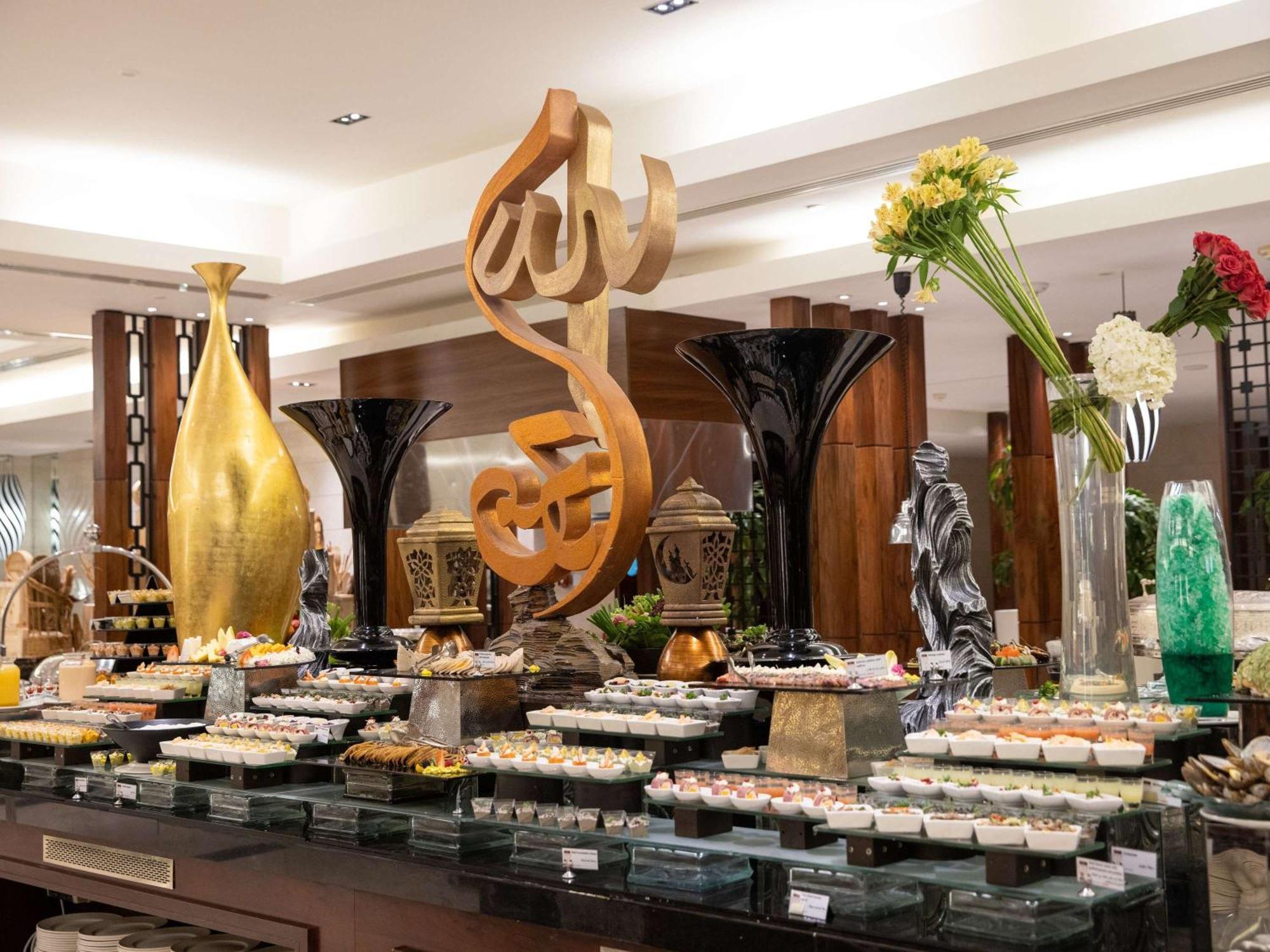 Swissotel Al Maqam Makkah Mekka Eksteriør billede The photo shows a beautifully arranged buffet display in a restaurant. In the center, there is a large decorative piece featuring Arabic calligraphy. Surrounding it are various elegant food presentations, including trays of desserts, likely reflectin