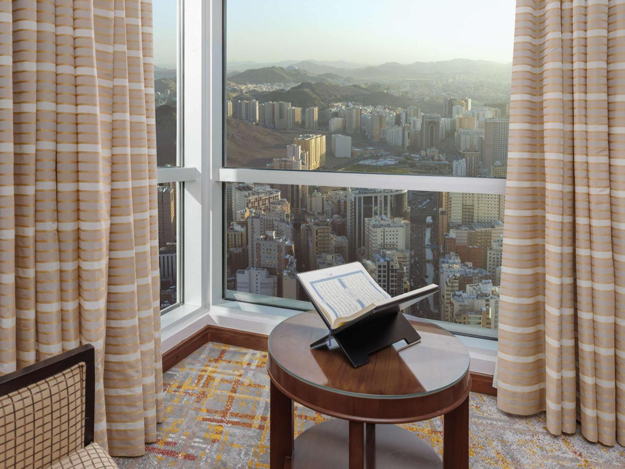 Swissotel Al Maqam Makkah Mekka Eksteriør billede The photo shows a corner of a hotel room with a large window offering a view of a cityscape in the background. Sunlight is filtering through the window, creating a bright atmosphere. Inside the room, there is a round wooden table with an open book or