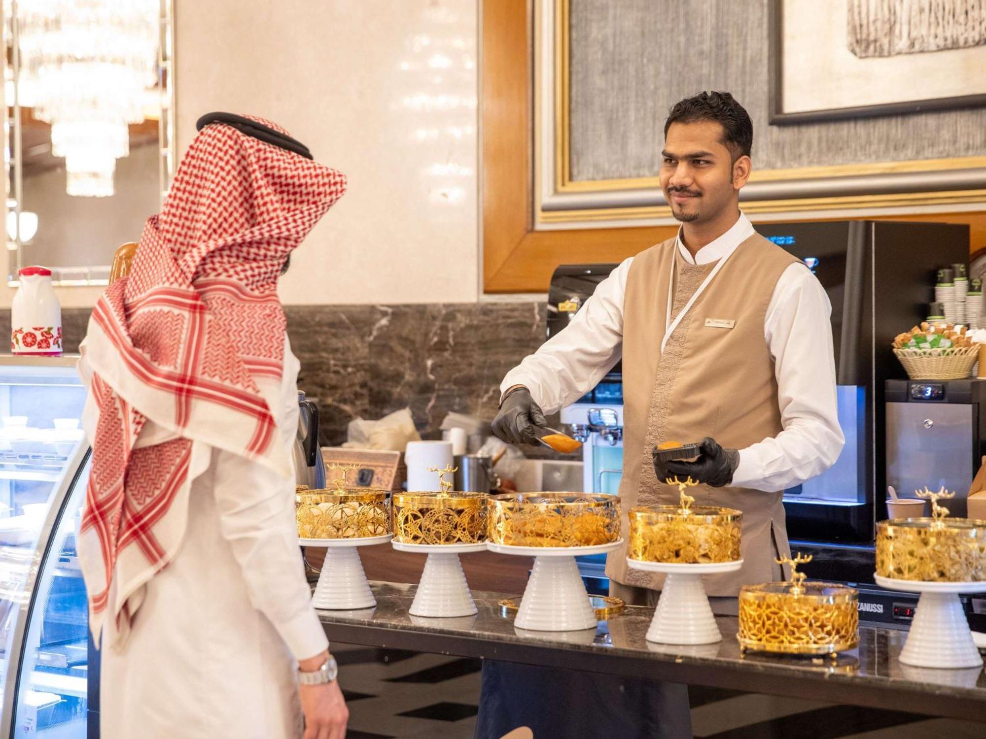 Swissotel Al Maqam Makkah Mekka Eksteriør billede The photo depicts a scene in a bakery or dessert shop. There is a display of various beautifully decorated cakes arranged on stands. A man wearing traditional attire, possibly a thobe and a headscarf, is interacting with a staff member who is smiling