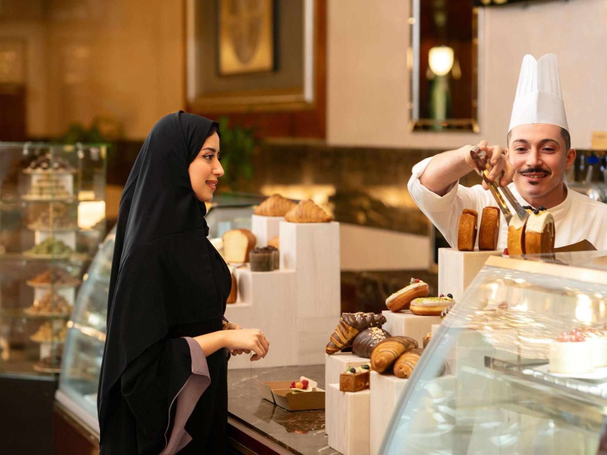Swissotel Al Maqam Makkah Mekka Eksteriør billede The photo shows a bakery or pastry shop scene. On the left, a woman is wearing a black abaya and appears to be interacting with a male baker. The baker, dressed in a white chef's hat and uniform, is behind a display counter filled with various baked 