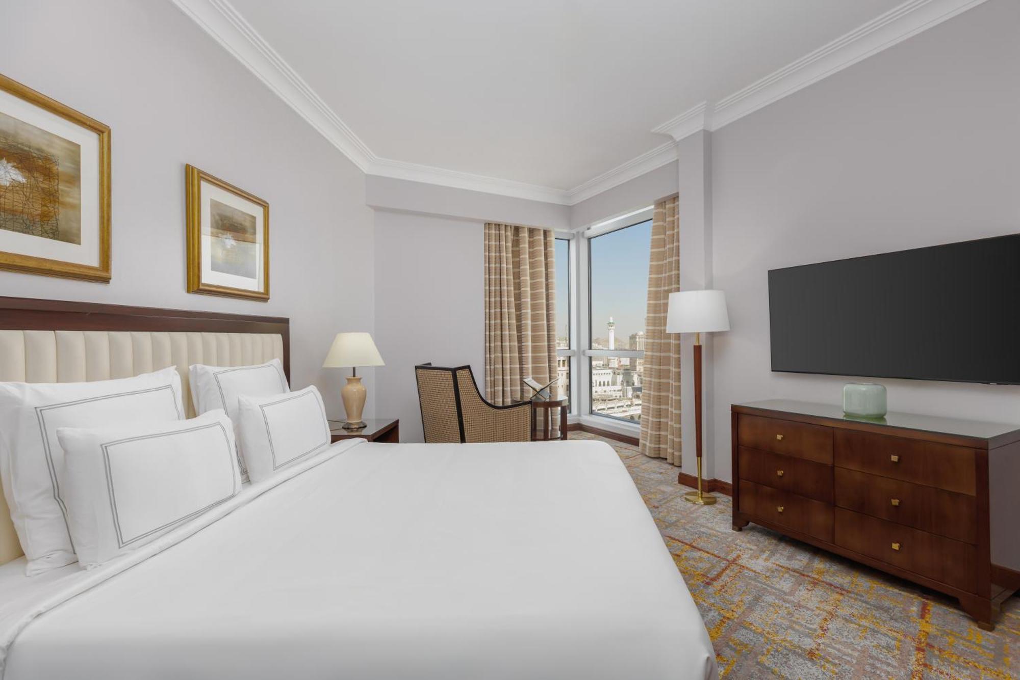 Swissotel Al Maqam Makkah Mekka Eksteriør billede The photo shows a hotel room featuring a neatly made bed with white linens and several decorative pillows. There's a comfortable chair positioned near a window, which has light curtains allowing natural light to enter. A bedside lamp and framed artwo